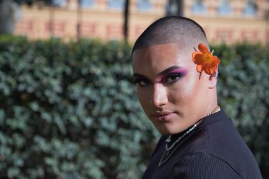 İkili olmayan genç ve Güney Amerikalı 'nın portresi, ağır makyajlı, kulağına güzel bir çiçek konmuş ve yeşillikle çevrili. Kavram kraliçesi, lgbtq +, gurur, nonoş, çiçekler.
