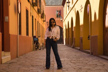 Beyaz düğümlü gömlekli, kot pantolonlu ve güneş gözlüklü genç ve güzel Güney Amerikalı kadın kaldırım taşlı bir caddenin ortasında poz veriyor. Güzellik, moda, tatil, seyahat, şehir, şehir kavramları.