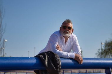 Olgun, gri saçlı, sakallı, güneş gözlüklü, beyaz gömlekli, deri ceketli, ufka endişeli bir şekilde bakan bir adam. Olgun erkek kavramı, emeklilik, işsizlik, sorunlar, yalnızlık.