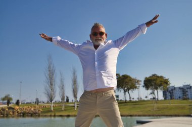 Olgun bir adam, gri saçlı, sakallı, güneş gözlüklü, beyaz gömlekli ve açık kahverengi pantolonlu, göl kenarında mutlu bir şekilde havaya atlıyor. Mutluluk kavramı, neşe, atlama, tatil, emeklilik.