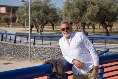 Olgun, sakallı, güneş gözlüklü, beyaz gömlekli, deri ceketli, dışarıda korkuluklara yaslanmış gri saçlı bir adam. Büyükbaba, zengin baba, yaşam tarzı, olgun adam.