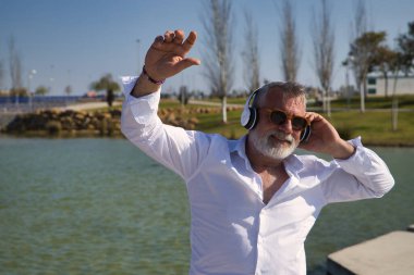 Olgun, sakallı, güneş gözlüklü, beyaz gömlekli, beyaz kulaklıklı, göl kenarında müzik dinlerken mutlu bir şekilde dans eden bir adam. Mutluluk kavramı, neşe, müzik, disko, dans, emeklilik.