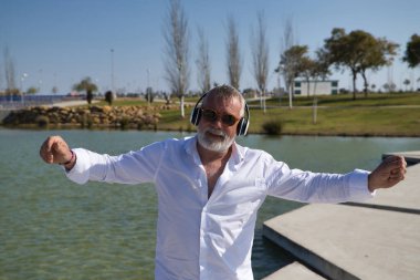 Olgun, sakallı, güneş gözlüklü, beyaz gömlekli, beyaz kulaklıklı, göl kenarında müzik dinlerken mutlu bir şekilde dans eden bir adam. Mutluluk kavramı, neşe, müzik, disko, dans, emeklilik.