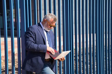 Olgun bir adam, yönetici, gri saçlı, sakallı, güneş gözlüklü, ceketli ve kravatlı, ofisin çıkışındaki laptoptan e-postaları kontrol eden. Yönetici kavramı, iş adamı, iş adamı, uygulamalar, dizüstü bilgisayar.