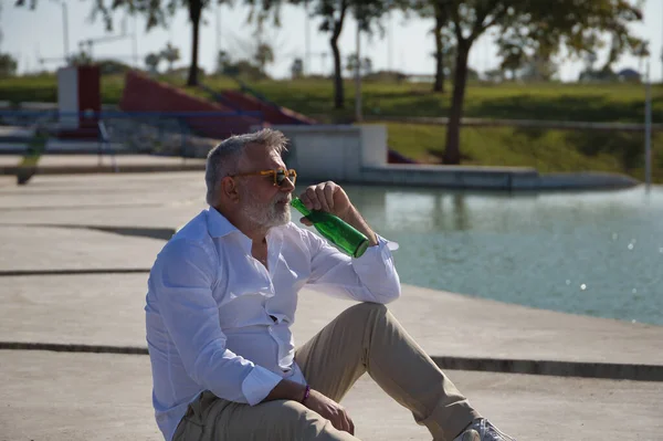 Olgun, sakallı, güneş gözlüklü, beyaz gömlekli, açık kahverengi pantolonlu, ağzında bir şişe birayla gölün yanında oturan bir adam. Olgun erkek kavramı, alkol, içki, tatil, sorunlar.