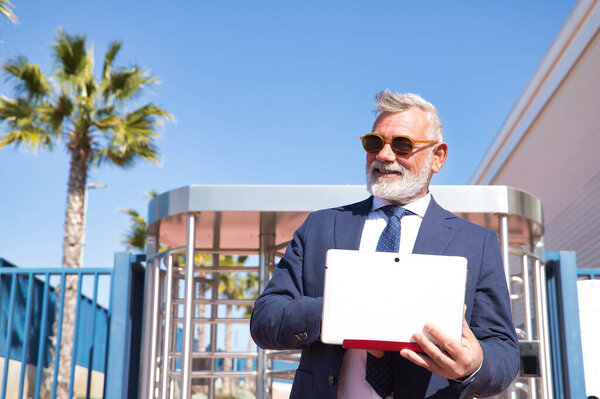 Mature man, professor, gray haired, bearded, with sunglasses, jacket and tie, checking emails on the laptop at the exit of the university. Rector concept, businessman, business, applications, laptop.