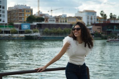 Genç ve güzel bir kadın, esmer, kıvırcık saçlı, güzel bir gülüşü olan, beyaz bir gömlek, kot pantolon ve güneş gözlüğü giyen nehir kenarındaki korkuluğa dayanan. Güzelliği, modayı, modeli, eğilimi, mutluluğu kavra.