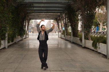 Sakallı, at kuyruklu, siyah tişörtlü, beyaz benekli, siyah pantolonlu ve ceketli, şehrin ortasında flamenko dansı yapan genç bir adam. Sanat, dans, kültür, gelenek.