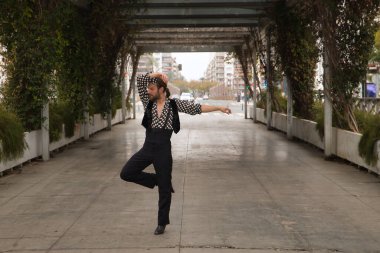 Sakallı, at kuyruklu, siyah tişörtlü, beyaz benekli, siyah pantolonlu ve ceketli, şehrin ortasında flamenko dansı yapan genç bir adam. Sanat, dans, kültür, gelenek.