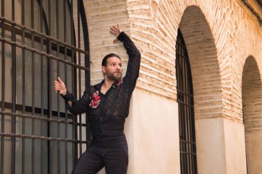 Sakallı, at kuyruklu, siyah şeffaf tişörtlü, siyah puantiyeli, kırmızı güllü, siyah pantolonlu ve ceketli, pencere ızgarasına yapışmış flamenko dansı yapan genç bir adam. Sanat kavramı, dans..