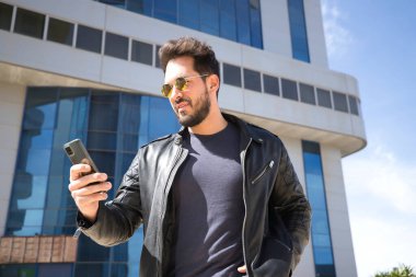 Sakallı, güneş gözlüklü, deri ceketli, cep telefonundan sosyal ağları kontrol eden yakışıklı bir genç. Güzelliği, modayı, eğilimi, uygulamayı, akıllı telefonu, sosyal ağları kavra.