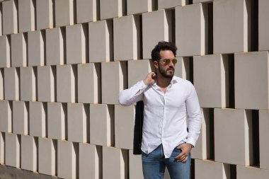 Sakallı, güneş gözlüklü, deri ceketli, beyaz gömlekli ve kot pantolonlu, beyaz taş bloklardan bir duvarın yanında yakışıklı bir genç. Güzelliği, modayı, eğilimi kavra.