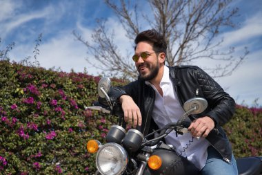 Sakallı, güneş gözlüklü, deri ceketli, beyaz gömlekli ve kot pantolonlu, büyük yer değiştiren motosikletli yakışıklı bir genç. Güzellik, moda, trend, motor, motosiklet, çıplak kavram.