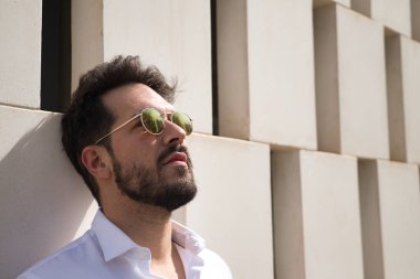 Sakallı, güneş gözlüklü ve beyaz gömlekli yakışıklı bir adamın portresi. Kafası beyaz taş bir duvara dayanıyor. Güzelliği, modayı, trendi, güneşi, yalnızlığı kavra.