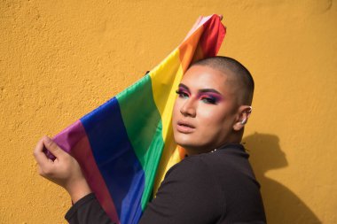Portrait of non-binary person, young and South American, heavily make up and holding a gay pride flag in a sensual way. Concept queen, lgbtq+, pride, queer.