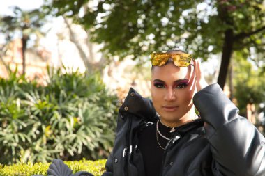 İkili olmayan genç ve Güney Amerikalı birinin portresi. Makyajlı ve kafasında aynalı güneş gözlüğü var. Etrafı yeşillikle çevrili. Kavram kraliçesi, lgbtq +, gurur, nonoş.