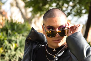 Portrait of non-binary person, young and South American, heavily make up, looking over mirrored sunglasses and surrounded by greenery. Concept queen, lgbtq+, pride, queer.