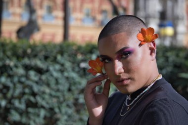 İkili olmayan genç ve Güney Amerikalı 'nın portresi, ağır makyajlı, kulağına güzel bir çiçek konmuş ve yeşillikle çevrili. Kavram kraliçesi, lgbtq +, gurur, nonoş, çiçekler.