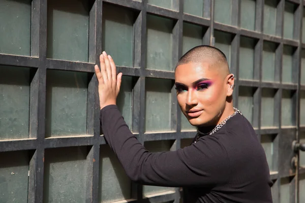 Portrait of non-binary person, young and South American, heavily make up, leaning against a wall, receiving the sun rays. Concept queen, lgbtq+, pride, queer.