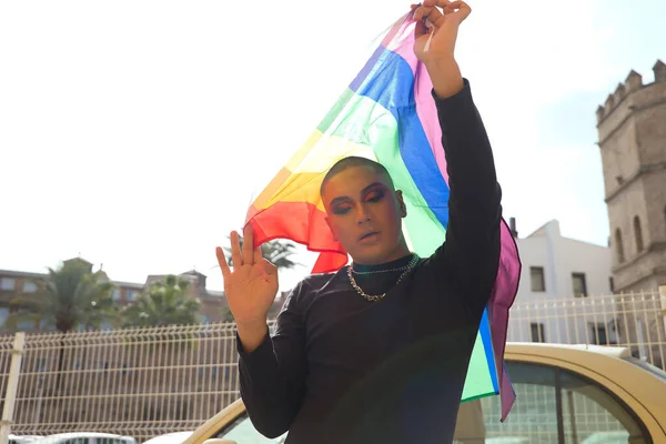 Portrait of non-binary person, young and South American, heavily make up and holding a gay pride flag with strong backlight. Concept queen, lgbtq+, pride, queer.