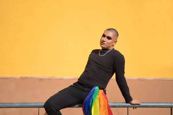 Non-binary person, young South American, heavily make up, sitting on a railing with a gay pride flag held by a trouser pocket. Concept queen, lgbtq+, pride, queer.