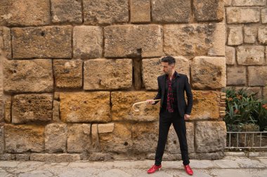 Siyah gömlekli, ceketli ve pantolonlu, kırmızı puantiyeli siyah atkı, kırmızı ayakkabılı ve bastonlu taş bir duvarın yanında duran genç bir İspanyol. Flamenko konsepti, sanat, dans, kültür, gelenek.