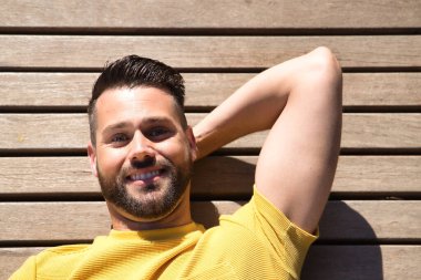 Yakışıklı genç bir adamın portresi, mavi gözlü ve mükemmel gülüşlü, sarı tişörtlü, tahta bir yüzeye uzanmış mutlu bir şekilde gülümseyen. Güzelliği, modayı, mutluluğu, eğilimi kavra.