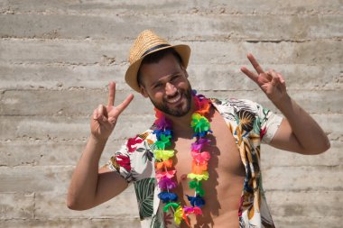 Genç ve yakışıklı bir adam, mavi gözlü, mükemmel gülüşlü, sakallı, şapkalı, Hawaii gömlekli ve çiçek kolyeli gri arka planda zafer işareti yapıyor. Tatiller, partiler, seyahatler.