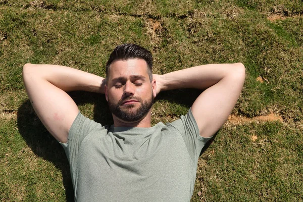 Young and handsome man, with a beard and green shirt, with his eyes closed and his hands behind the back of his neck lying on the grass, resting. Concept beauty, fashion, trend, sleep.