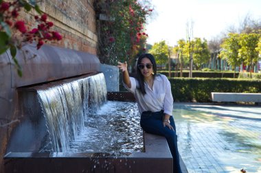 Genç ve güzel Güney Amerikalı kadın, güneş gözlüklü ve beyaz gömlekli, Avrupa 'da tatilde, çeşmenin yanında oturuyor, eliyle su atıyor. Tatil, seyahat, güzellik, moda, su.