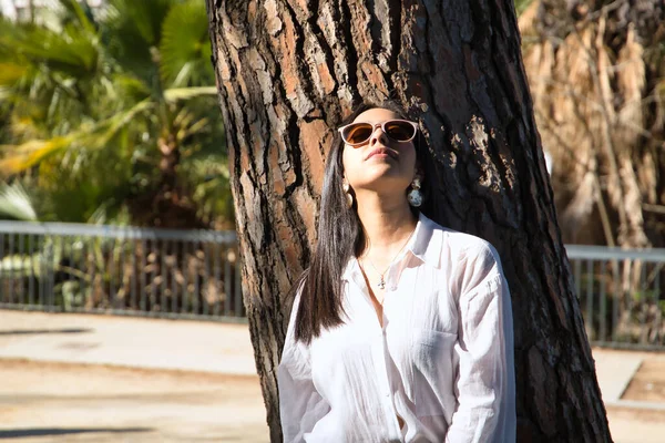 若くて美しい女性 南アメリカ サングラスと白いシャツを着て 木の幹に太陽の光を浴びて傾いている コンセプト旅行 美しさ ファッション — ストック写真