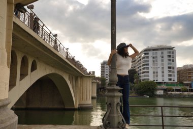 Genç ve güzel bir kadın, esmer, kıvırcık saçlı, beyaz gömlekli, kot pantolonlu ve şapkanın güneşliğinde bir el olan, yukarı ve bir sokak lambasına tutunan. Güzelliği, modayı, modeli, trendi, bulutları kavra.
