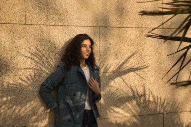 Genç ve güzel bir kadın, esmer, kıvırcık saçlı, ceketli ve dalgın bakışlı, palmiye yapraklarının arasında bir duvara yaslanmış. Güzelliği, modayı, modayı, modeli, sonbaharı, düşünceleri kavra.