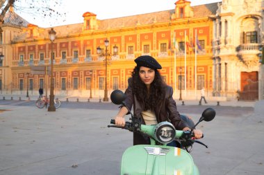 Genç ve güzel bir kadının portresi, esmer, kıvırcık saçlı, deri ceketli ve siyah bereli, şehre gelirken kiralık bir motosiklete monte edilmiş. Güzelliği, modayı, ulaşımı kavra.