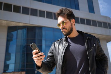 Sakallı, güneş gözlüklü, deri ceketli, cep telefonundan sosyal ağları kontrol eden yakışıklı bir genç. Güzelliği, modayı, eğilimi, uygulamayı, akıllı telefonu, sosyal ağları kavra.