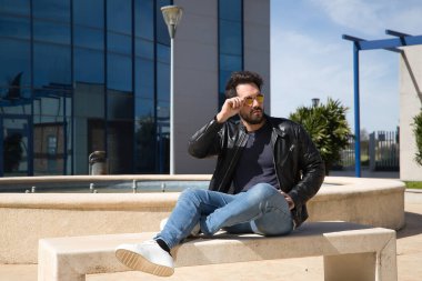 Sakallı, güneş gözlüklü, deri ceketli ve kotlu taş bir bankta oturan yakışıklı genç adam, rahatlamış. Güzellik, moda, trend, modern, rahatla.