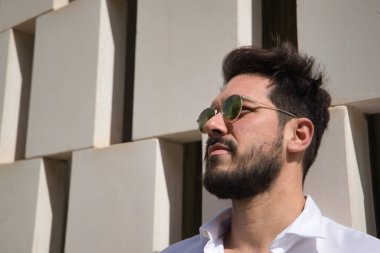 Sakallı, güneş gözlüklü ve beyaz gömlekli yakışıklı bir adamın portresi. Kafası beyaz taş bir duvara dayanıyor. Güzelliği, modayı, trendi, güneşi, yalnızlığı kavra.