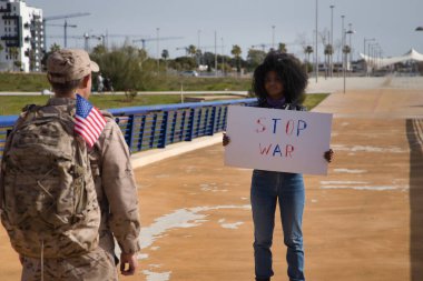 Savaşa hayır pankartıyla gelen Afro-Amerikalı kadın, ortağının önünde, göreve gitmek zorunda olan bir Amerikan askeri. Ordu, misyonlar, savaş, ordu. Kadınlara seçici odaklanma.