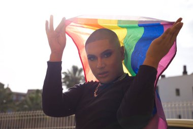 İkili olmayan genç ve Güney Amerikalı 'nın portresi ağır makyaj yapıyor ve arkasında güçlü bir ışık tutan bir gey gururu bayrağı taşıyor. Kavram kraliçesi, lgbtq +, gurur, nonoş.