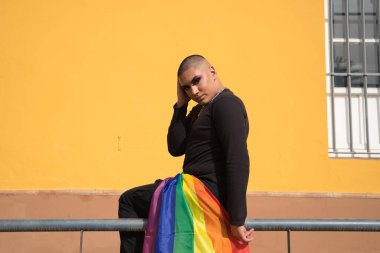 İkili olmayan, genç Güney Amerikalı, ağır makyajlı, pantolon cebinde bir gay gururu bayrağı taşıyan bir korkulukta oturuyor. Kavram kraliçesi, lgbtq +, gurur, nonoş.