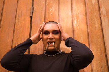İkili olmayan bir kişinin portresi, genç ve Güney Amerikalı, arka planda tahta bir duvar bulunan dişli metal bir zinciri ısırıyor. Kavram kraliçesi, lgbtq +, gurur, nonoş.