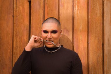 İkili olmayan bir kişinin portresi, genç ve Güney Amerikalı, arka planda tahta bir duvar bulunan dişli metal bir zinciri ısırıyor. Kavram kraliçesi, lgbtq +, gurur, nonoş.