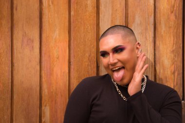 İkili olmayan bir kişinin portresi, genç ve Güney Amerikalı, ağır makyajlı, yüzünde bir el ve arka planda ahşap bir duvarla dilini uzatıyor. Kavram kraliçesi, lgbtq +, gurur, nonoş.
