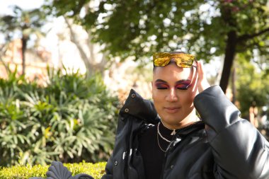 İkili olmayan genç ve Güney Amerikalı 'nın portresi, ağır makyajlı, gözleri kapalı ve kafasında aynalı güneş gözlüğü olan ve yeşilliklerle çevrili. Kavram kraliçesi, lgbtq +, gurur, nonoş.