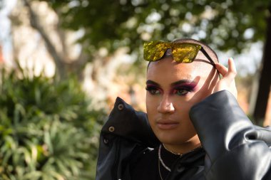 İkili olmayan birinin portresi, genç ve Güney Amerikalı, aşırı makyajlı, düşünceli ve kafasında aynalı güneş gözlükleriyle ve yeşilliklerle çevrili. Kavram kraliçesi, lgbtq +, gurur, nonoş.