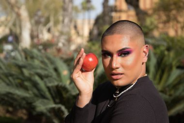 İkili olmayan bir kişinin portresi, genç ve Güney Amerikalı, ağır makyajlı, elinde kırmızı bir elma tutuyor ve bitki örtüsüyle çevrili. Konsept kraliçesi, lgbtq +, gurur, ibne, günah.