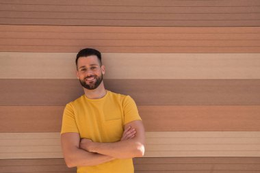 Mavi gözlü, mükemmel gülüşlü, sarı tişörtlü, kollarını kavuşturmuş ve mutlu bir şekilde gülümseyen yakışıklı bir genç adam portresi. Güzelliği, modayı, gülümsemeyi, mutluluğu, eğilimi kavra.