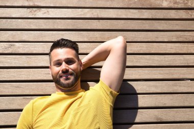 Yakışıklı genç bir adamın portresi, mavi gözlü ve mükemmel gülüşlü, sarı tişörtlü, tahta bir yüzeye uzanmış mutlu bir şekilde gülümseyen. Güzelliği, modayı, mutluluğu, eğilimi kavra.