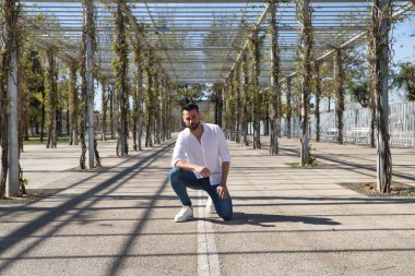 Sakallı, beyaz gömlekli, kot pantolonlu, dizlerinin üzerinde ve elleri bacaklarının üzerinde yakışıklı bir genç. Konsept moda, güzellik, modern moda, model.