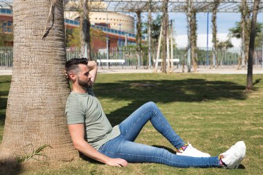 Yakışıklı genç adam, mavi gözlü, sakallı, yeşil tişörtlü ve kot pantolonlu, bir palmiye ağacının gövdesine yaslanmış, dinleniyor. Güzelliği, modayı, trendi, uykuyu, rüyayı kavra.